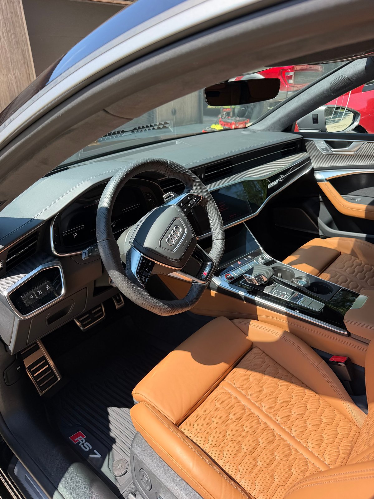 Interior of luxury car with orange leather seats, steering wheel, dashboard, and windshield view of red car outside